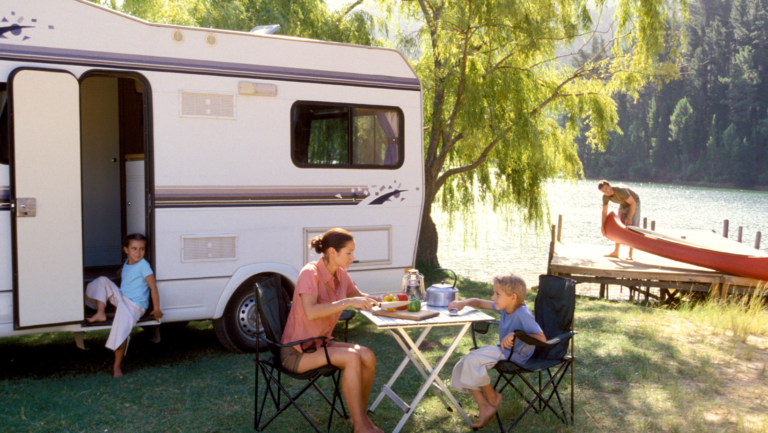 Familie beim Camping am See: Kinder sitzen am Wohnwagen und am Picknicktisch, Vater bereitet ein Kanu vor